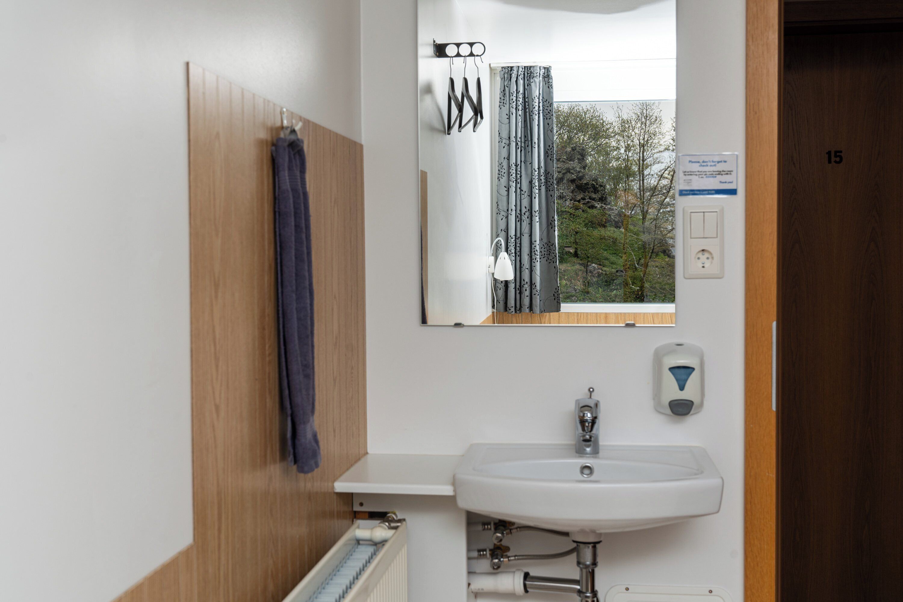 A bathroom with a wooden panel wall, a white sink, a mirror, a towel rack, and a window with a view of trees.