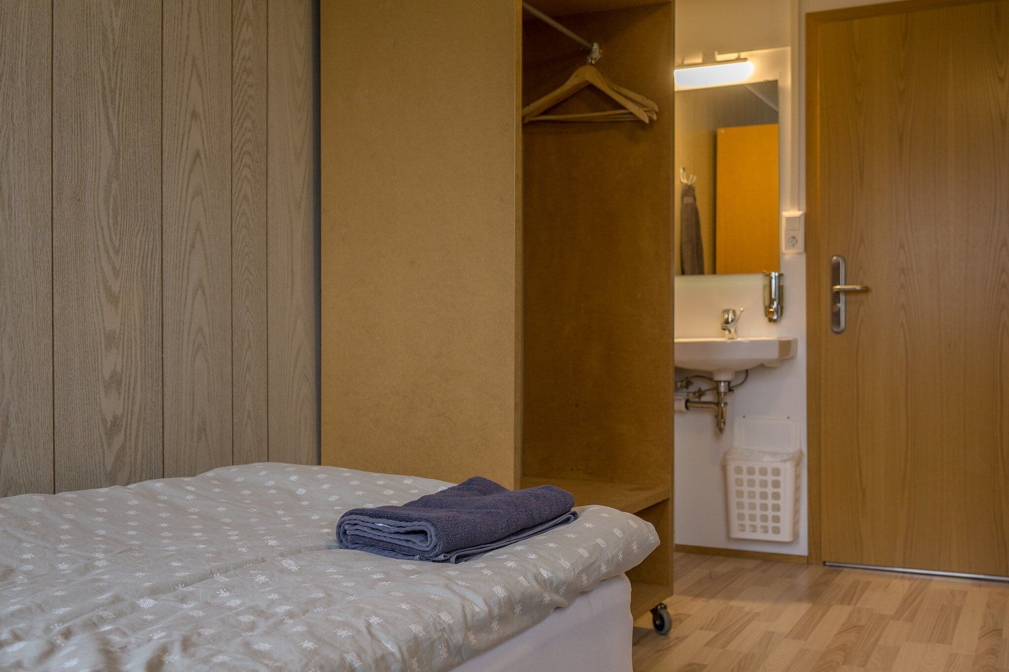 Un dormitorio con cama, perchero, baño con lavamanos y espejo, y una puerta de madera.