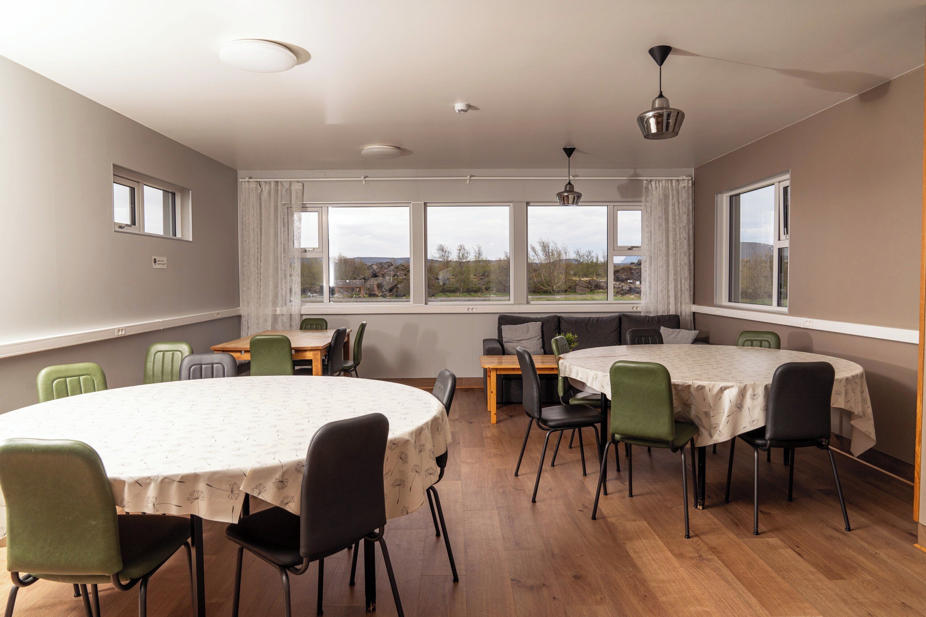 A dining area with round tables and chairs, large windows with curtains, and a view of the outdoors.