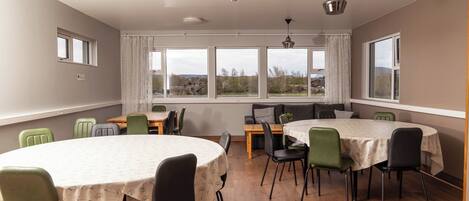 A dining area with round tables and chairs, large windows with curtains, and a view of the outdoors.