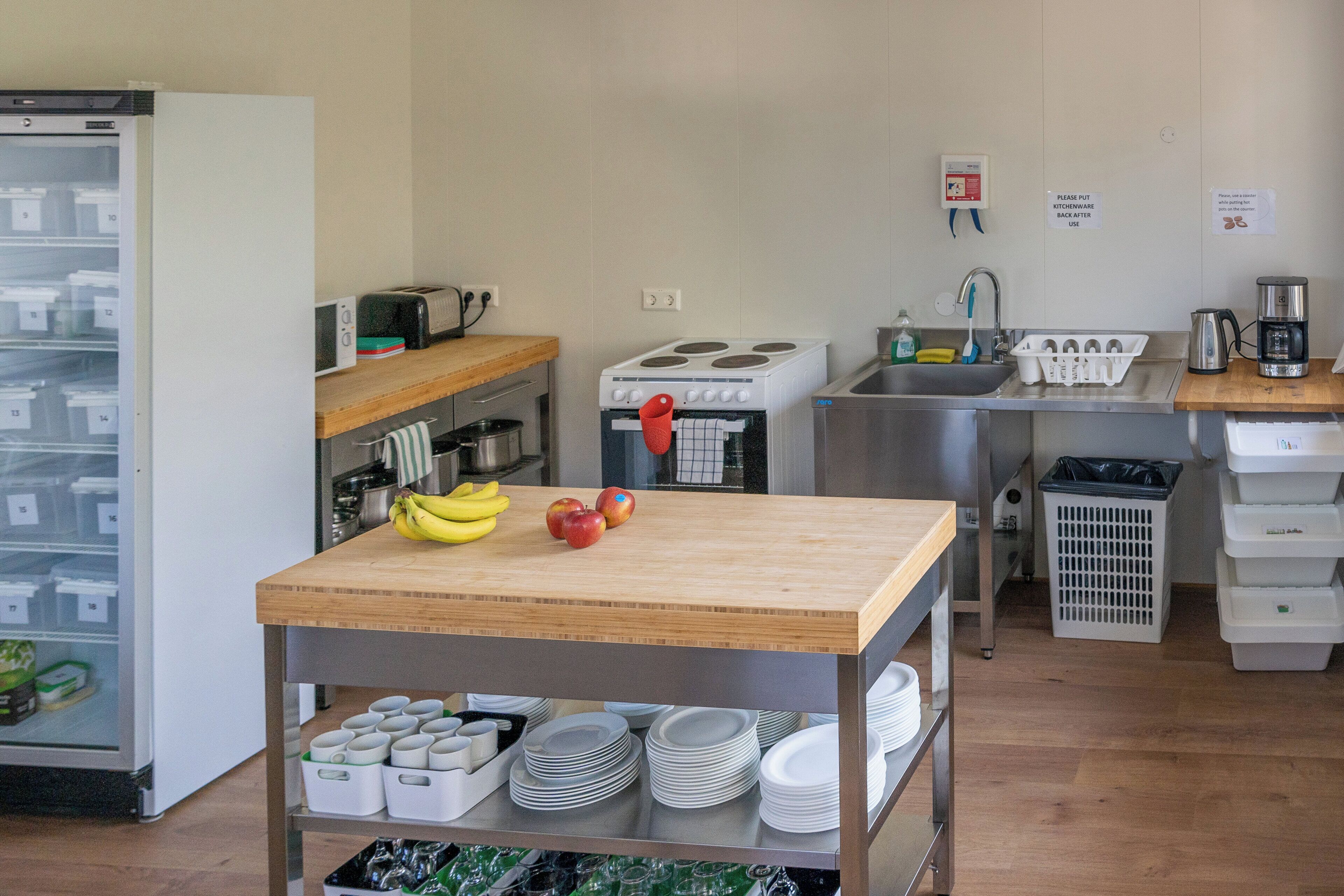 Een keuken met een houten tafel, een fornuis, een spoelbak en diverse keukenapparaten. Op de tafel liggen bananen en appels, en er staat een koelkast op de achtergrond.