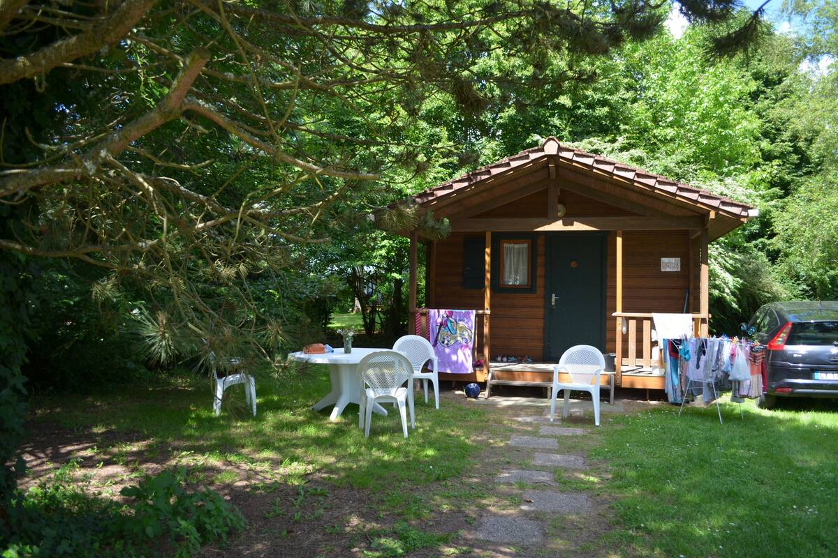 Eine kleine Holzhütte mit einer Veranda, umgeben von Bäumen. Ein Tisch mit Stühlen und ein Auto sind in der Nähe geparkt.