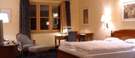 Quarto de hotel com cama, escrivaninha, cadeira, luminária de mesa, janela com cortinas e luminária na parede.