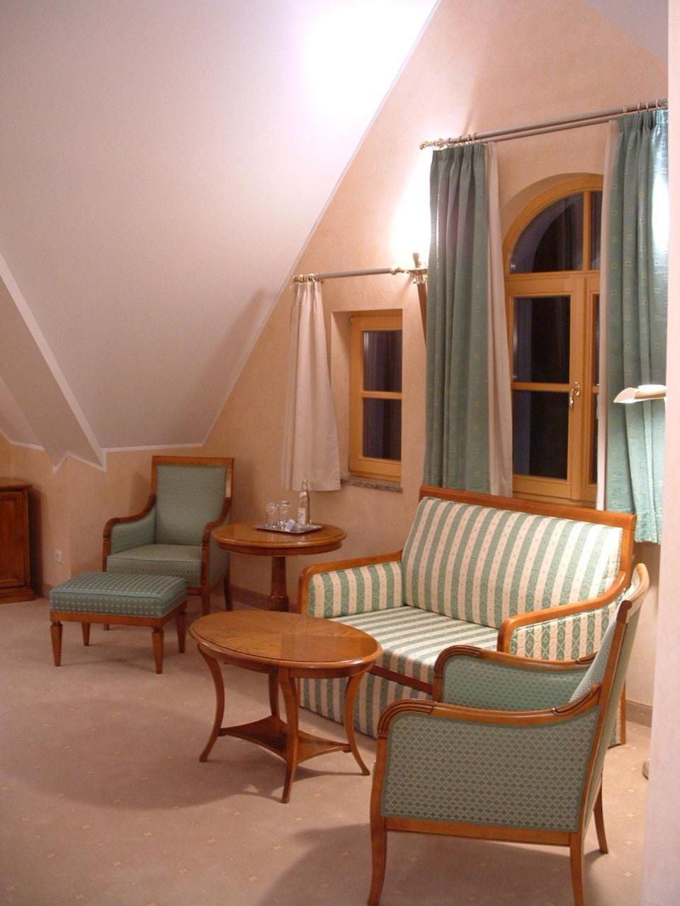 Una habitación acogedora con techo inclinado, un sofá, dos sillones, una mesita redonda y una ventana con cortinas.
