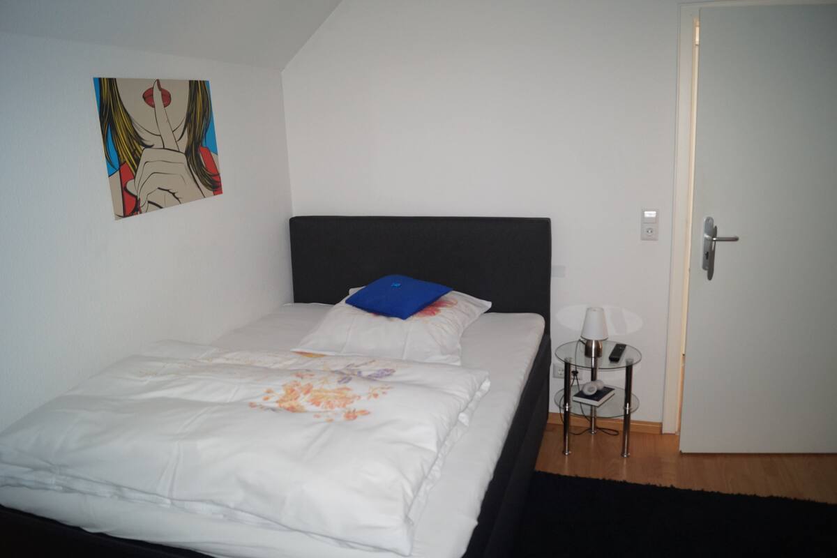 A neatly made bed with white bedding and a blue pillow, a small bedside table with a lamp, and a door to the right.