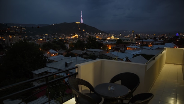 Un balcón con sillas y una mesa, con vistas a un paisaje urbano que incluye edificios y una torre distante.