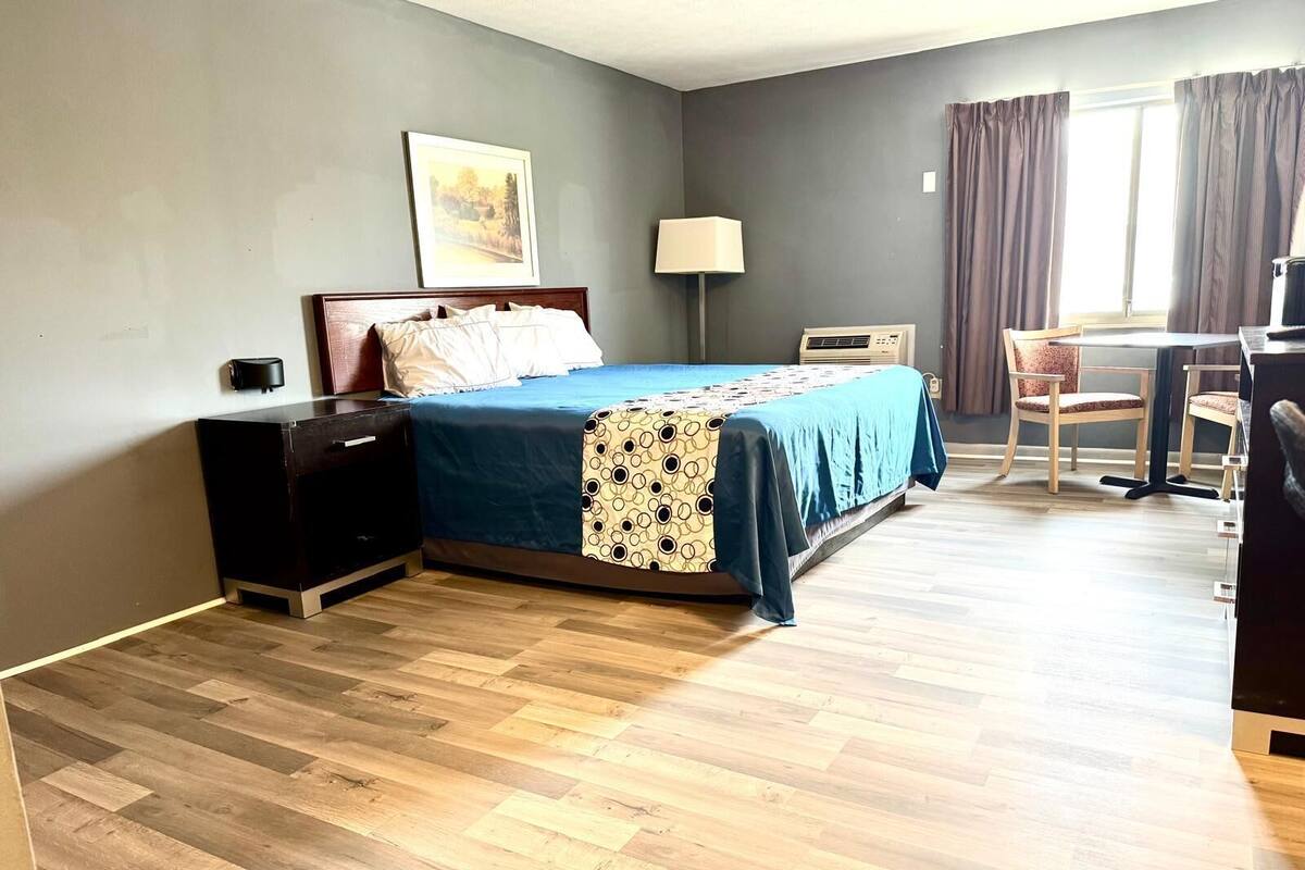 Quarto de hotel com cama, mesa de cabeceira, abajur, aparelho de ar condicionado e área de refeições com cadeiras.