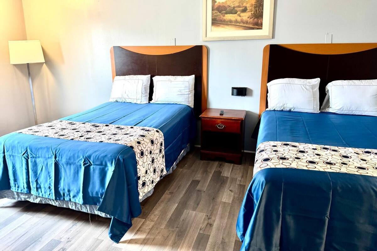 Quarto de hotel com duas camas, mesa de cabeceira, luminária e quadro na parede.