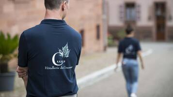 Una persona con una camisa azul oscuro que tiene un logo y texto en la parte trasera, caminando por una calle con edificios al fondo.