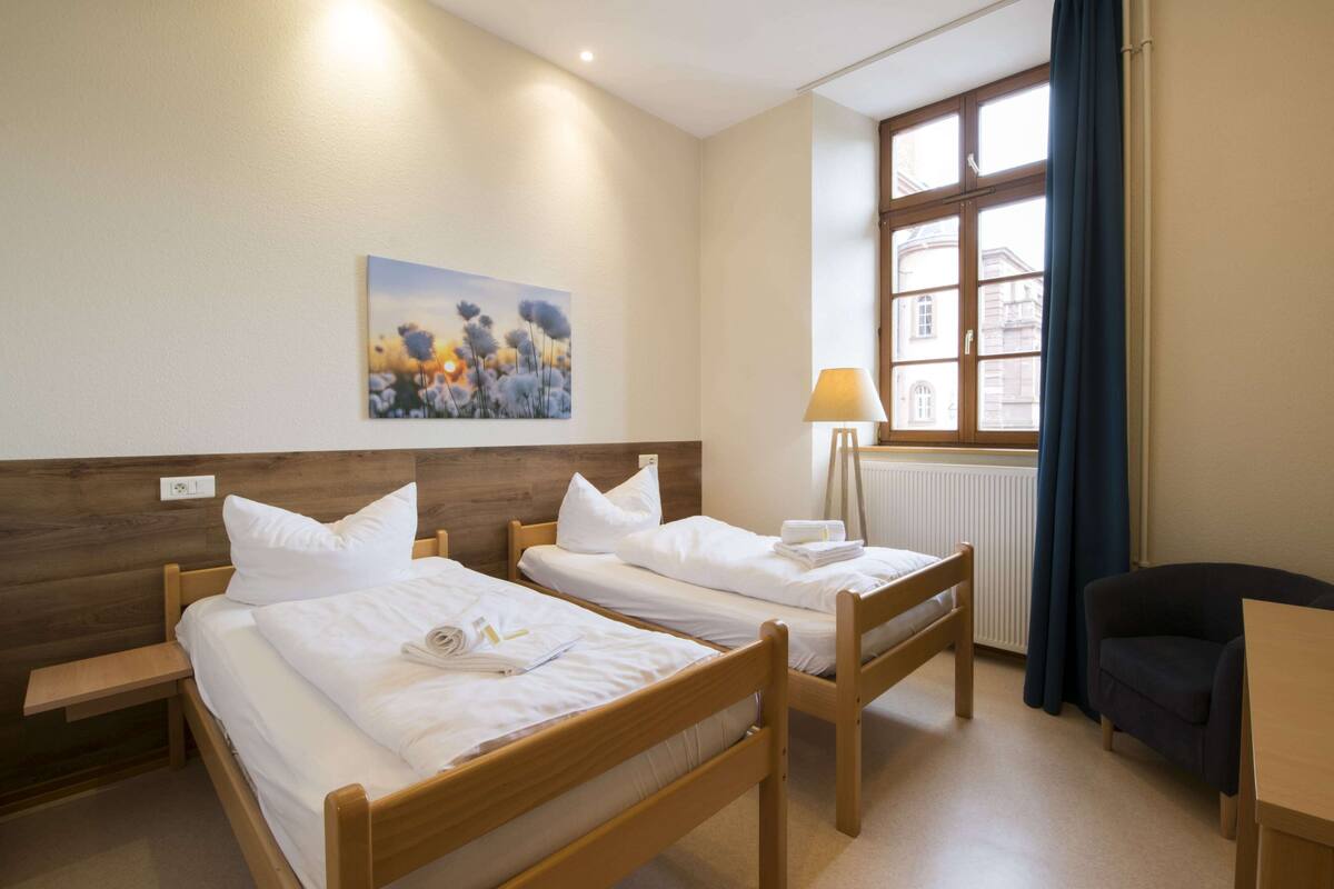 A hotel room with two wooden beds, a painting on the wall, a window with curtains, a lamp, and a chair.