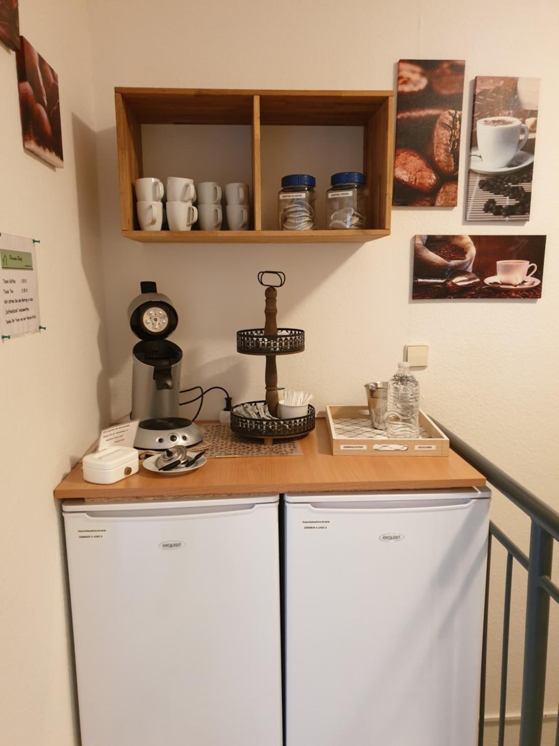 Een kitchenette met een koffieapparaat, waterkoker en een koffiemolen op een houten aanrecht. Daaronder staan twee koelkasten.