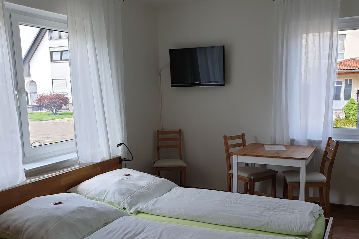A room with a bed, a TV mounted on the wall, a table with chairs, and a window with curtains.