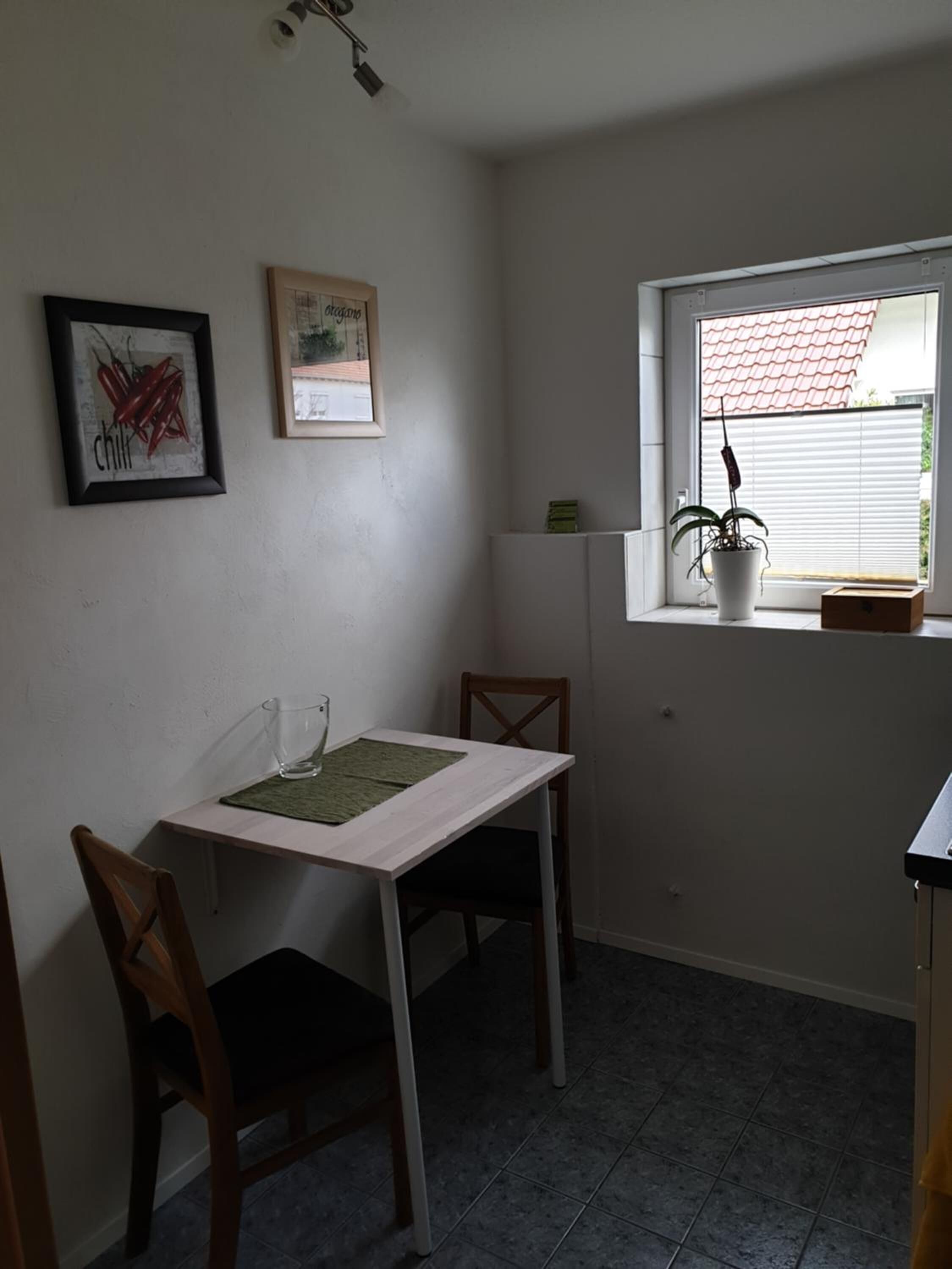 Ein kleiner, sauberer Raum mit einem weißen Tisch und zwei Stühlen, einem Fenster mit einer Pflanze und zwei gerahmten Bildern an der Wand.