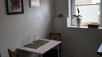 Ein kleiner, sauberer Raum mit einem weißen Tisch und zwei Stühlen, einem Fenster mit einer Pflanze und zwei gerahmten Bildern an der Wand.
