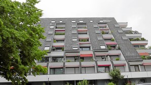 En modern byggnad med flera balkonger, varav några har planteringar, och en asfalterad markyta med parkerade bilar.