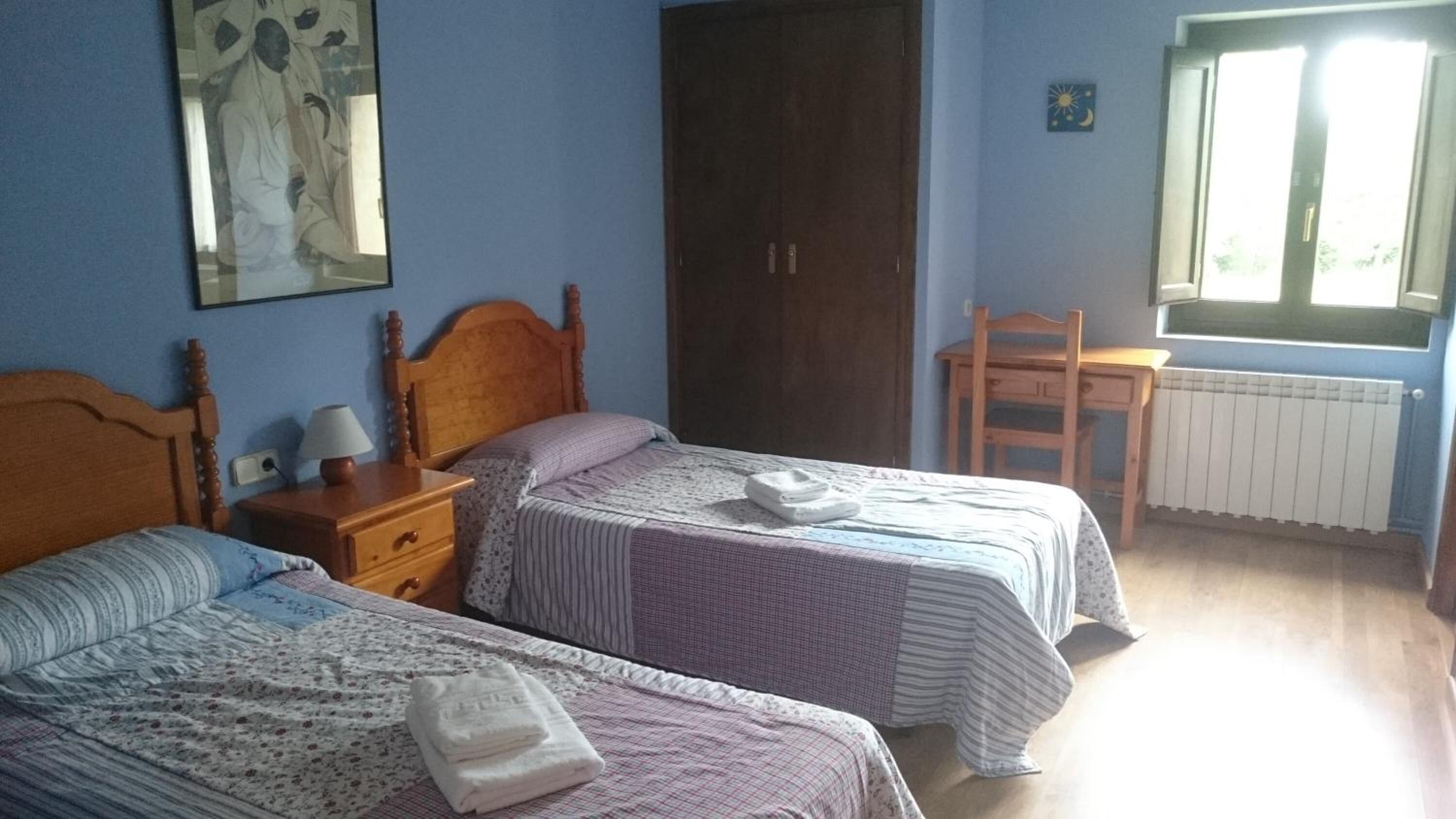 Un dormitorio con dos camas, una mesita de noche, una silla de madera, un radiador y una ventana con vistas al verdor.