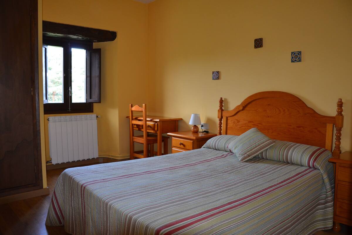 Une chambre à coucher avec un lit, une table de chevet en bois, un bureau avec une chaise, une fenêtre et un radiateur.
