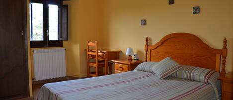 A bedroom with a bed, a wooden nightstand, a desk with a chair, a window, and a radiator.