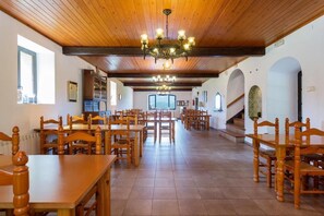 A spacious dining hall with wooden tables and chairs, a chandelier, and a view of the kitchen in the background. - Hostal Falgars (La Pobla de Lillet)