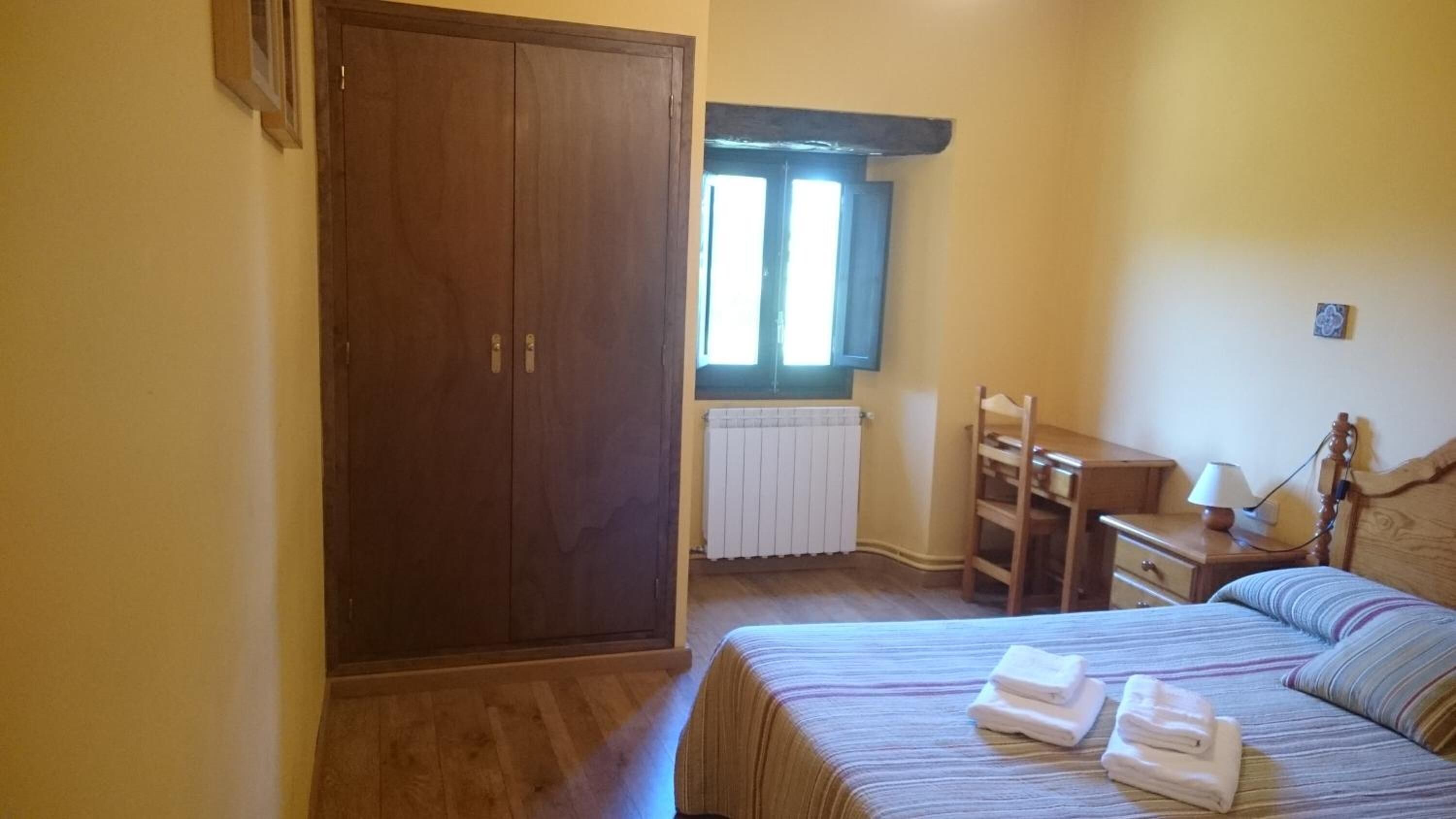 Un dormitorio con un armario de madera, una ventana con persianas, un radiador, una mesita de noche con una lámpara y una cama con toallas dobladas.