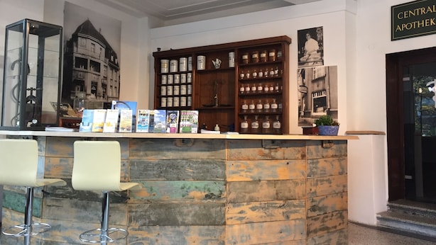 Un espace d’accueil avec un comptoir en pierre, deux tabourets de bar, un meuble en bois contenant des bocaux et une enseigne indiquant « Central Apotheke ».