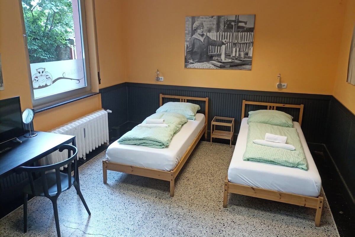 A room with two wooden beds, a desk with a chair, a radiator, and a window with a view of trees.