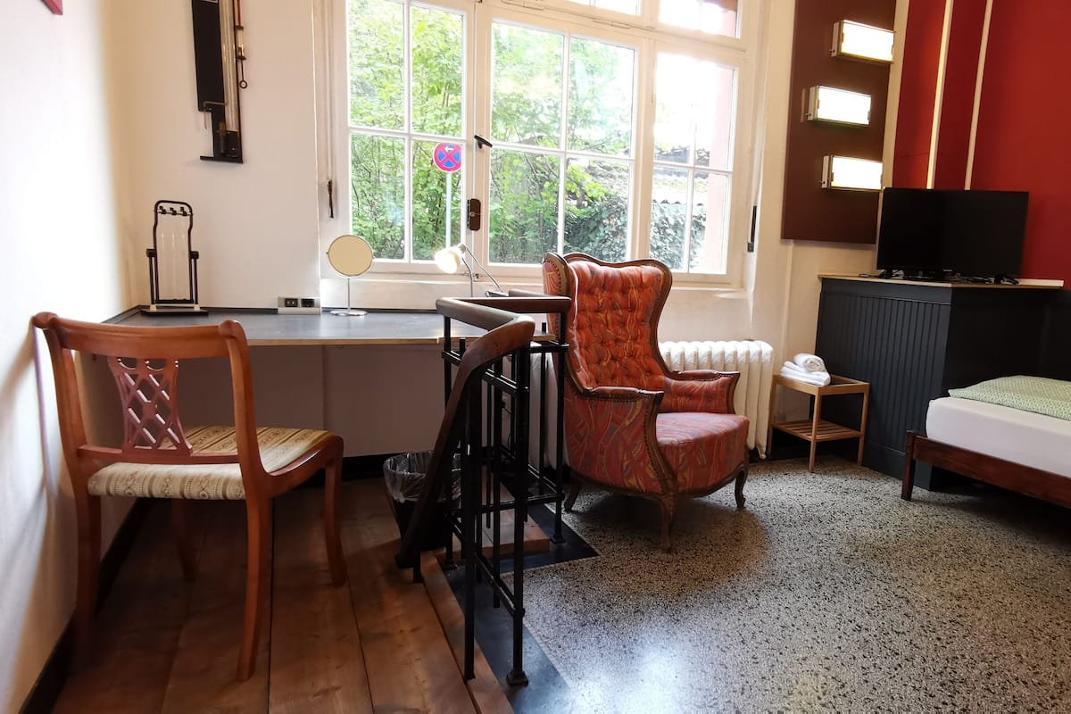 A room with a desk, two chairs, a bed, a radiator, a television, and a window with a view of trees.