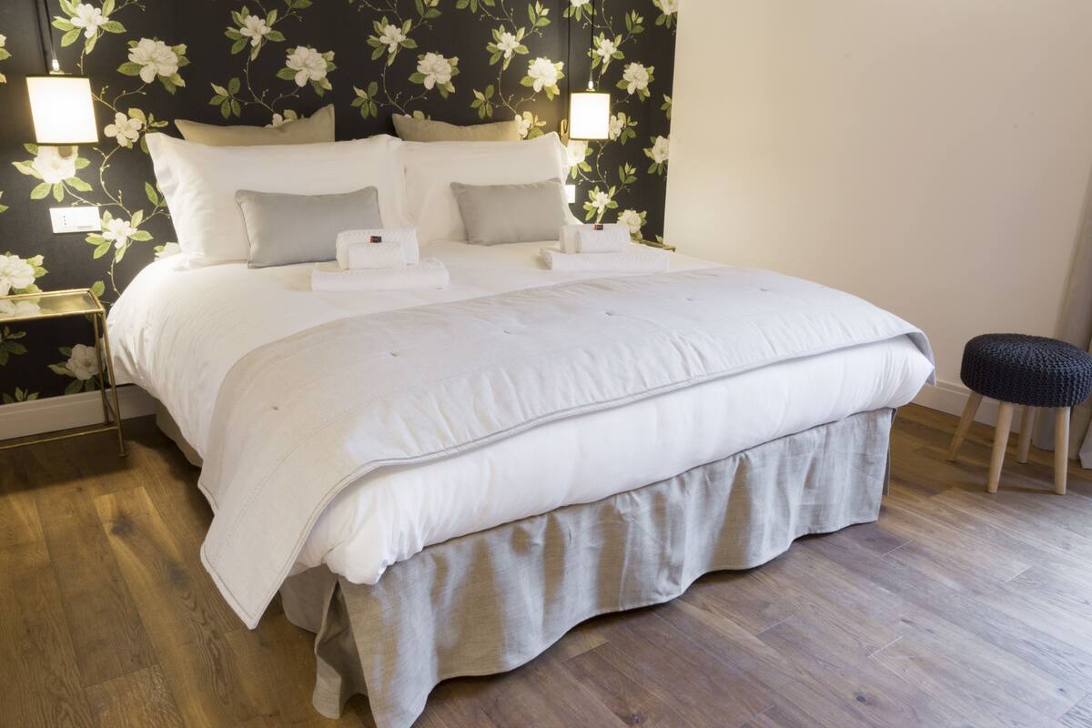 A neatly made bed with white linens and a grey skirt, a nightstand with a lamp, and a floral wallpaper pattern on the wall.