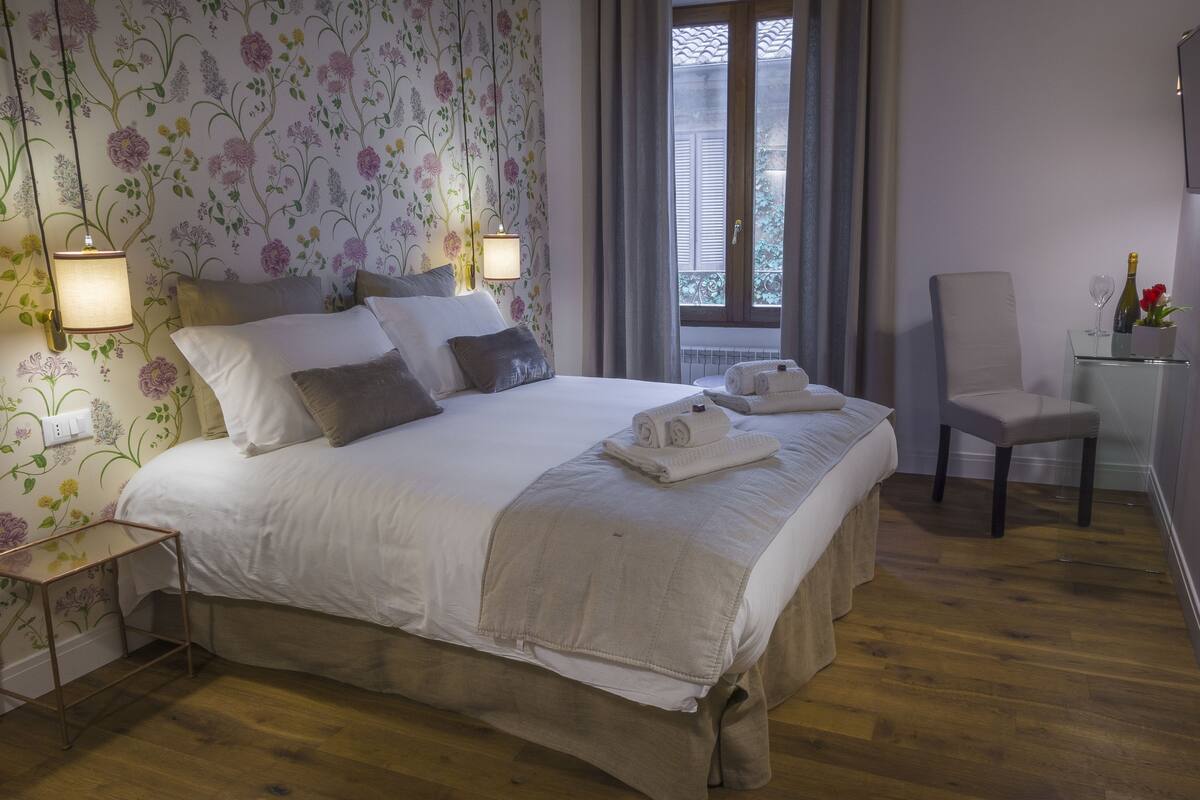 A neatly made bed with a white comforter and several pillows, a small bedside table with a lamp, a chair, and a window with curtains.