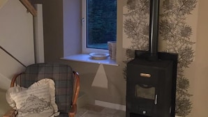 A cozy room with a stone floor, a plaid chair, a window, and a black wood-burning stove. - Dunruadh Cottage (Alexandria)