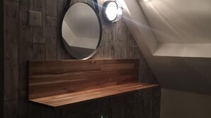A room with wooden panel walls, a round mirror, a wooden shelf, a stool, and a slanted ceiling with a light fixture. - Dunruadh Cottage (Alexandria)