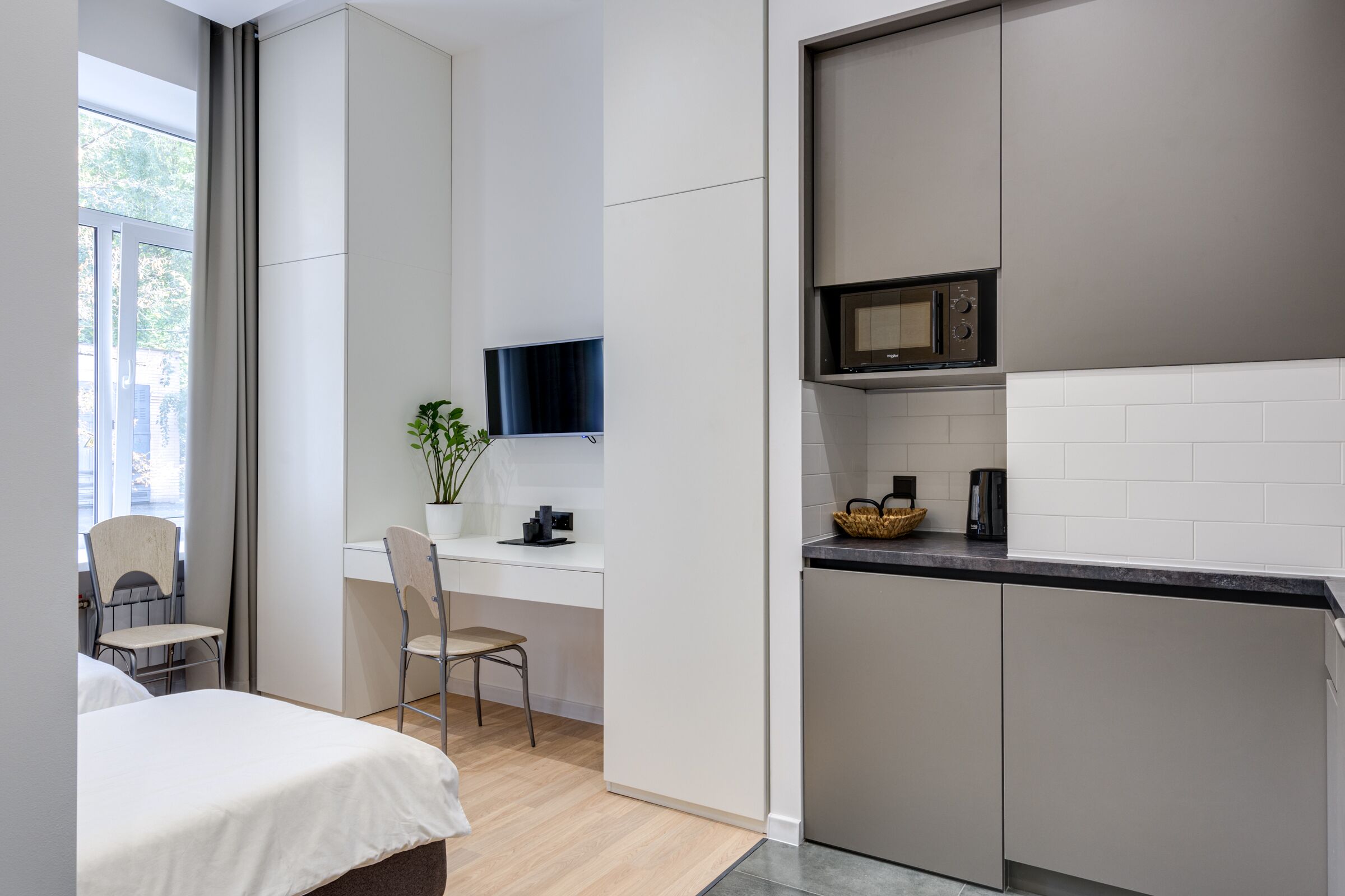 Ein gemütliches Zimmer mit Bett, Schreibtisch, Stuhl, Blumentopf, Fernseher und einer Küchenzeile mit Mikrowelle.