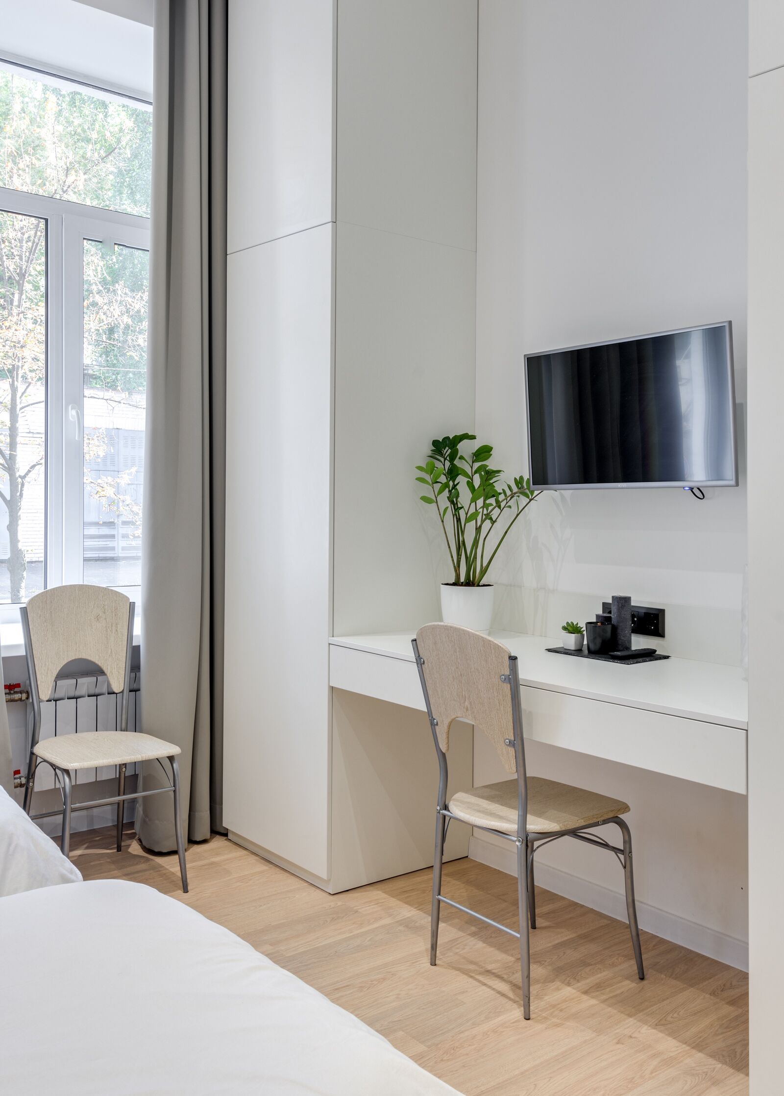 Habitación de hotel moderna con un escritorio blanco, dos sillas, un televisor de pantalla plana, una planta en maceta y una cama.