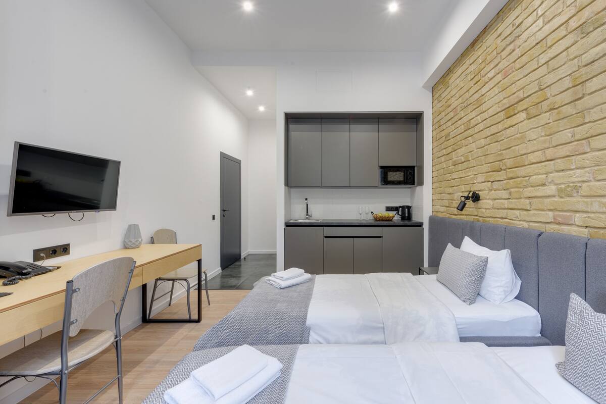 Quarto de hotel moderno com cama, escrivaninha, cadeira e área de cozinha com parede de tijolos.