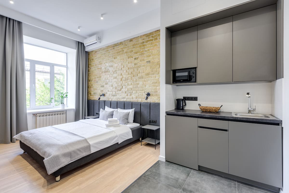 Quarto moderno com cama grande, mesa de cabeceira pequena, cozinha compacta com micro-ondas e janela com cortinas.