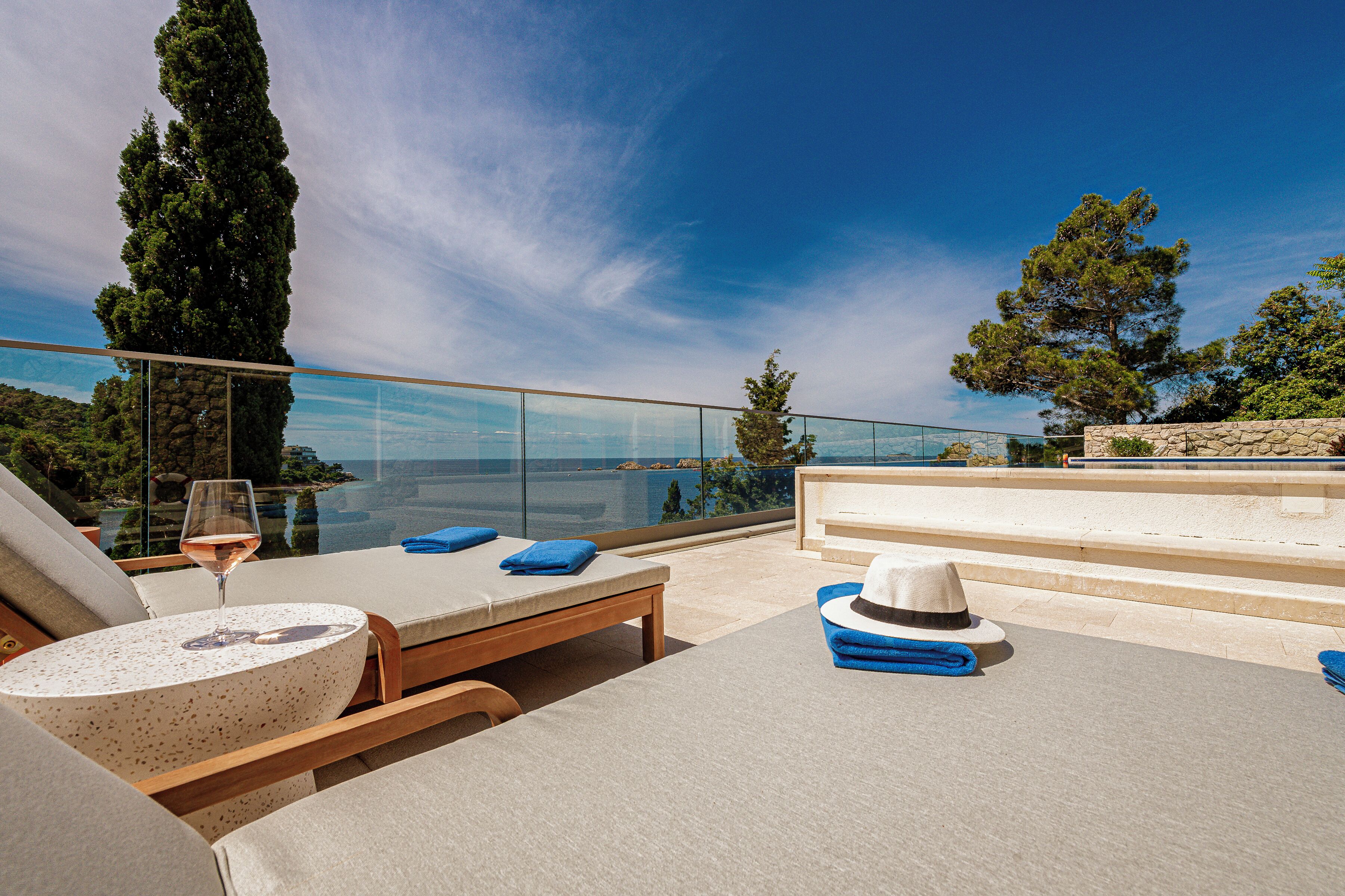 A sunlit outdoor lounge area with lounge chairs, a table with a glass of wine, and a view of the sea and greenery.