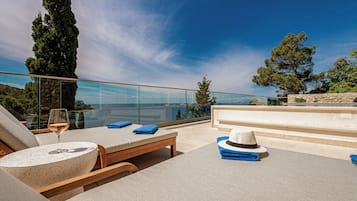 A sunlit outdoor lounge area with lounge chairs, a table with a glass of wine, and a view of the sea and greenery.