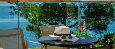 A round table with a book, a hat, and a glass of red wine, set against a scenic view of the sea and trees.