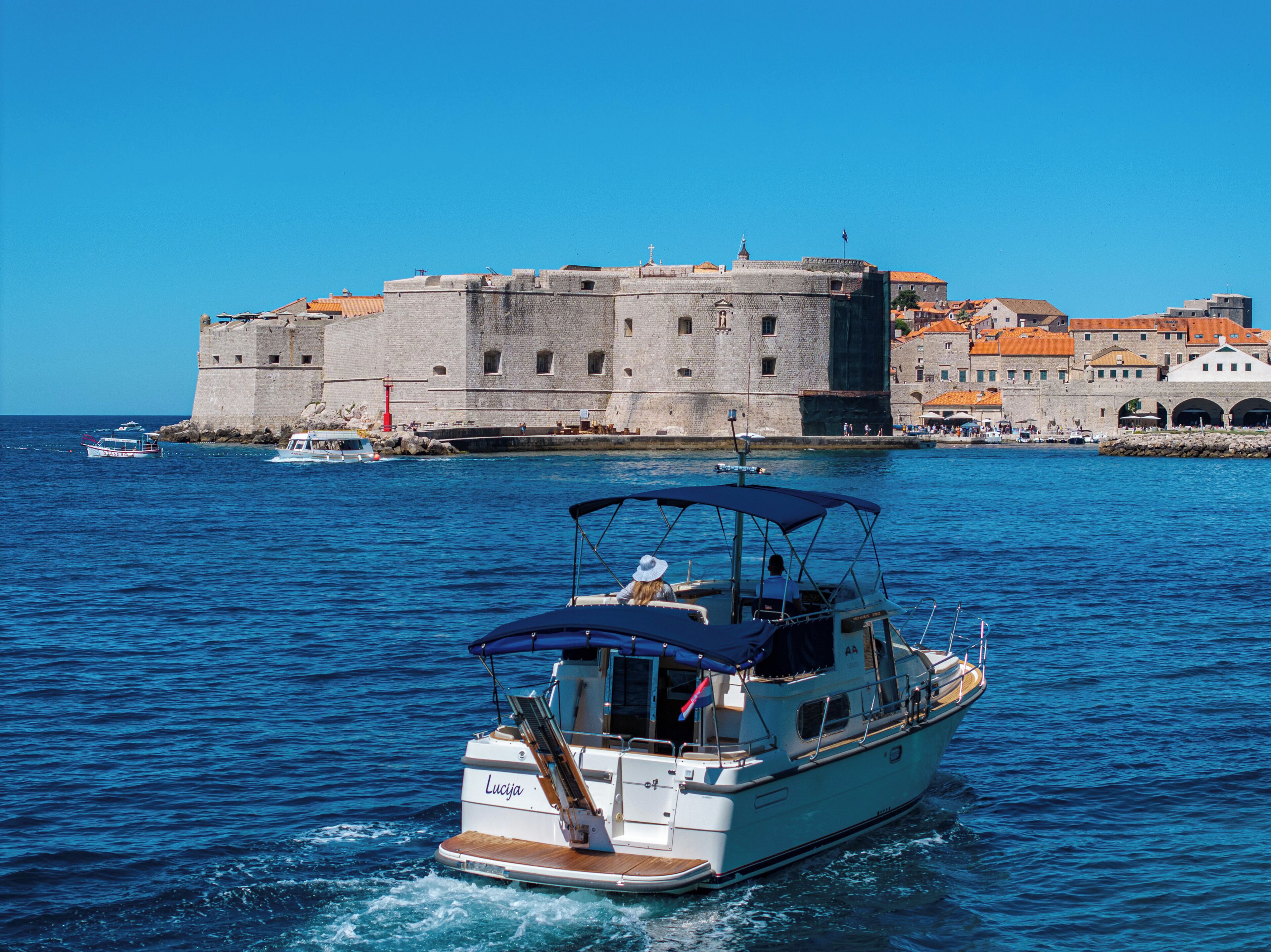 Un bateau appelé « Lucija », doté d’un pont couvert, naviguait à proximité d’une forteresse historique en pierre, composée de plusieurs bâtiments et surmontée d’un phare rouge.