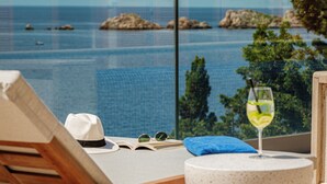 Una tumbona con sombrero, libro y una copa de bebida, con vista a un mar azul y despejado con rocas y árboles.