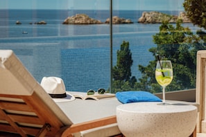 Un transat agrémenté d’un chapeau, d’un livre et d’un verre, offrant une vue imprenable sur une mer bleue et limpide, avec des rochers et des arbres.