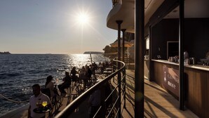 A sunlit outdoor dining area with tables and chairs, overlooking the sea, and a bar with a sign reading 'cave ba more'. - Villa Malo More (Dubrovnik)