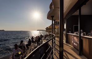 A sunlit outdoor dining area with tables and chairs, overlooking the sea, and a bar with a sign reading 'cave ba more'. - Villa Malo More (Dubrovnik)