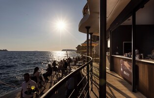 A sunlit outdoor dining area with tables and chairs, overlooking the sea, and a bar with a sign reading 'cave ba more'.
