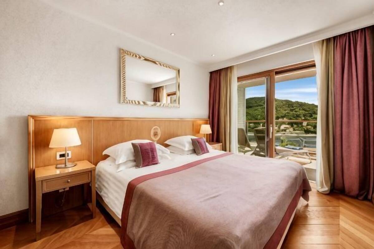 A hotel room with a large bed, a nightstand, a mirror, and a view of a mountainous landscape.