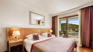 Habitación de hotel con una cama grande, una mesita de noche con lámpara, un espejo y vistas a un balcón con un paisaje pintoresco.