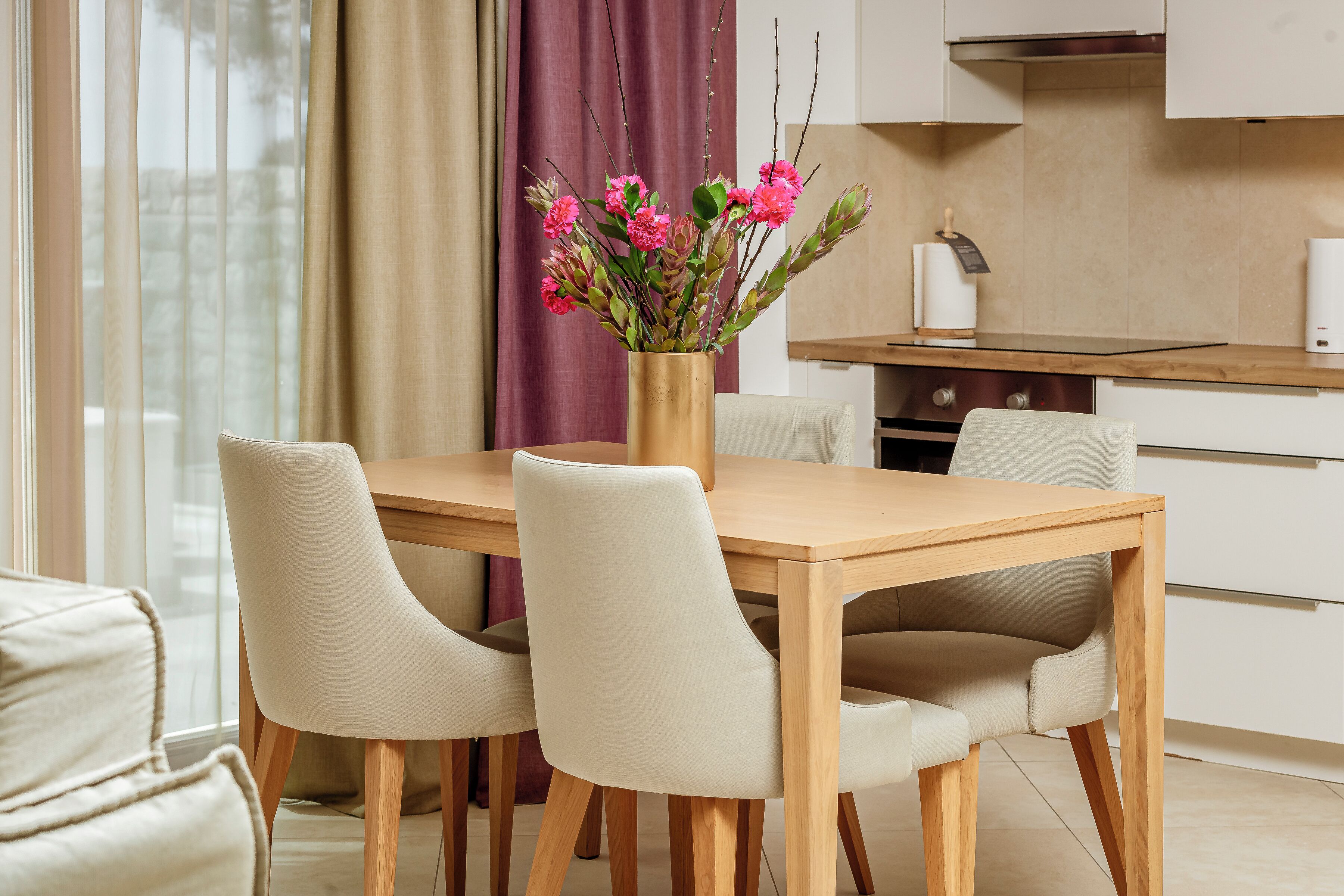Uma sala de jantar moderna com uma mesa de madeira e quatro cadeiras, um vaso com flores rosa e uma cozinha ao fundo.