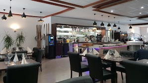 A restaurant with multiple dining tables, black chairs, and a bar area with a counter and various bottles.