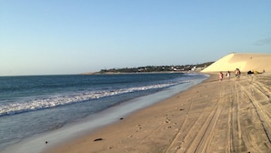 Una playa arenosa con huellas y surcos de neumáticos, cielo despejado y una duna distante con gente.