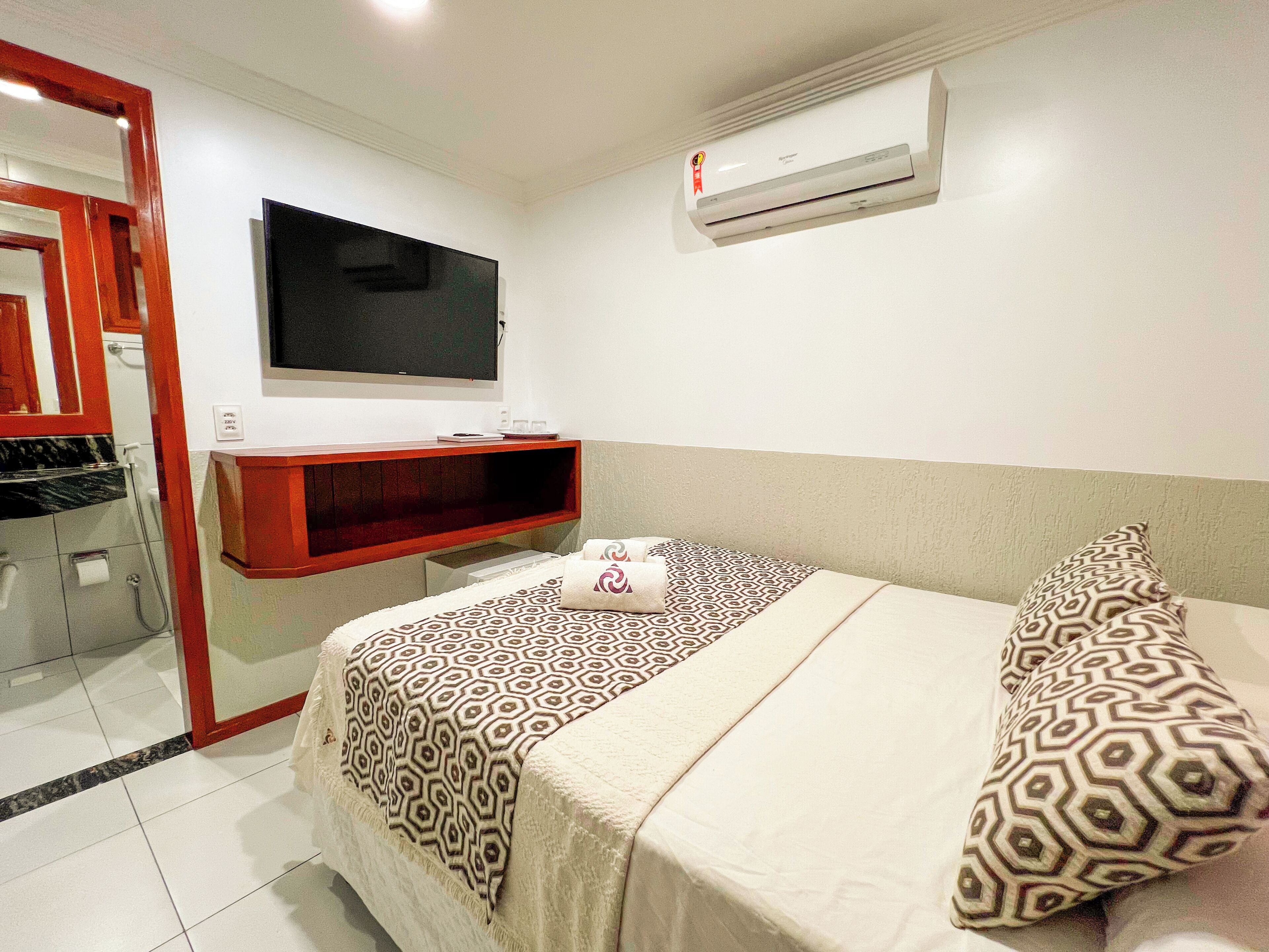 Quarto de hotel com cama, televisão, prateleira vermelha e área de duche.